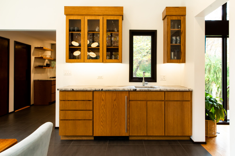 This is an example of a small midcentury single-wall wet bar in Chicago with an undermount sink, flat-panel cabinets, medium wood cabinets, granite benchtops, porcelain floors, grey floor and black benchtop.