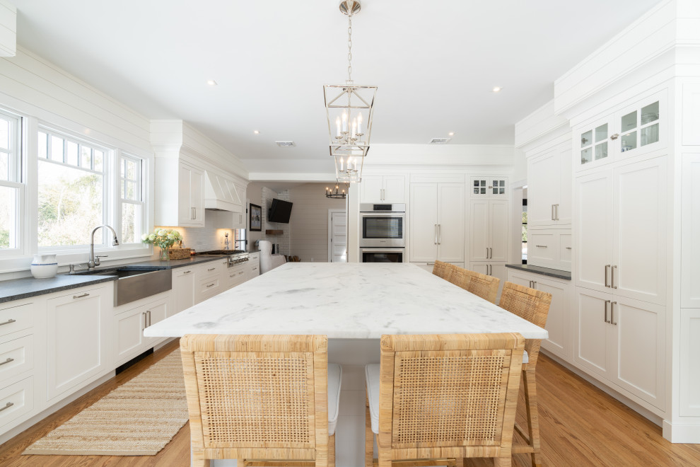 Expansive country galley eat-in kitchen in New York with a farmhouse sink, shaker cabinets, white cabinets, granite benchtops, white splashback, ceramic splashback, stainless steel appliances, medium hardwood floors, with island, brown floor and grey benchtop.