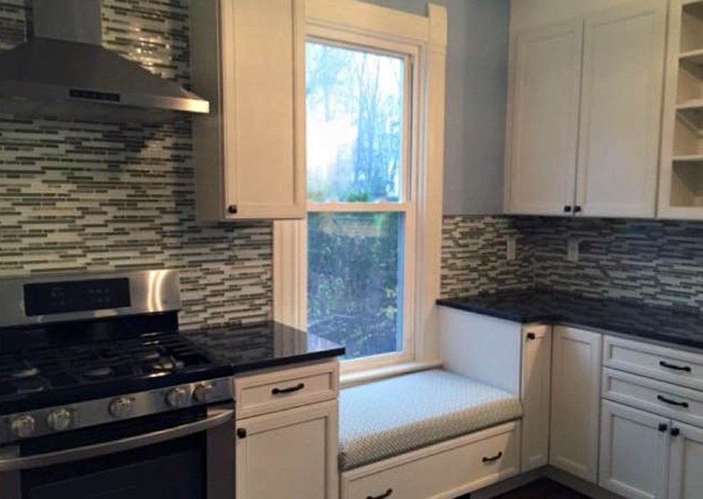 Example of a mid-sized transitional l-shaped medium tone wood floor and brown floor kitchen pantry design with a farmhouse sink, recessed-panel cabinets, white cabinets, granite countertops, multicolored backsplash, glass tile backsplash, stainless steel appliances, no island and black countertops