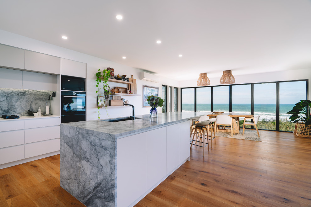 Photo of a large l-shaped eat-in kitchen in Sunshine Coast with a double-bowl sink, flat-panel cabinets, white cabinets, grey splashback, black appliances, medium hardwood floors, with island, brown floor and beige benchtop.