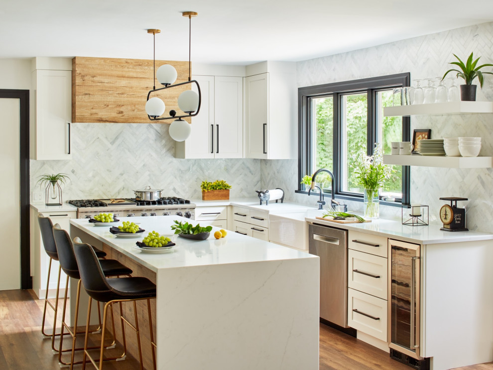 This is an example of a mid-sized contemporary u-shaped eat-in kitchen in New York with a farmhouse sink, shaker cabinets, white cabinets, white splashback, stainless steel appliances, with island, brown floor, white benchtop, stone tile splashback and laminate floors.