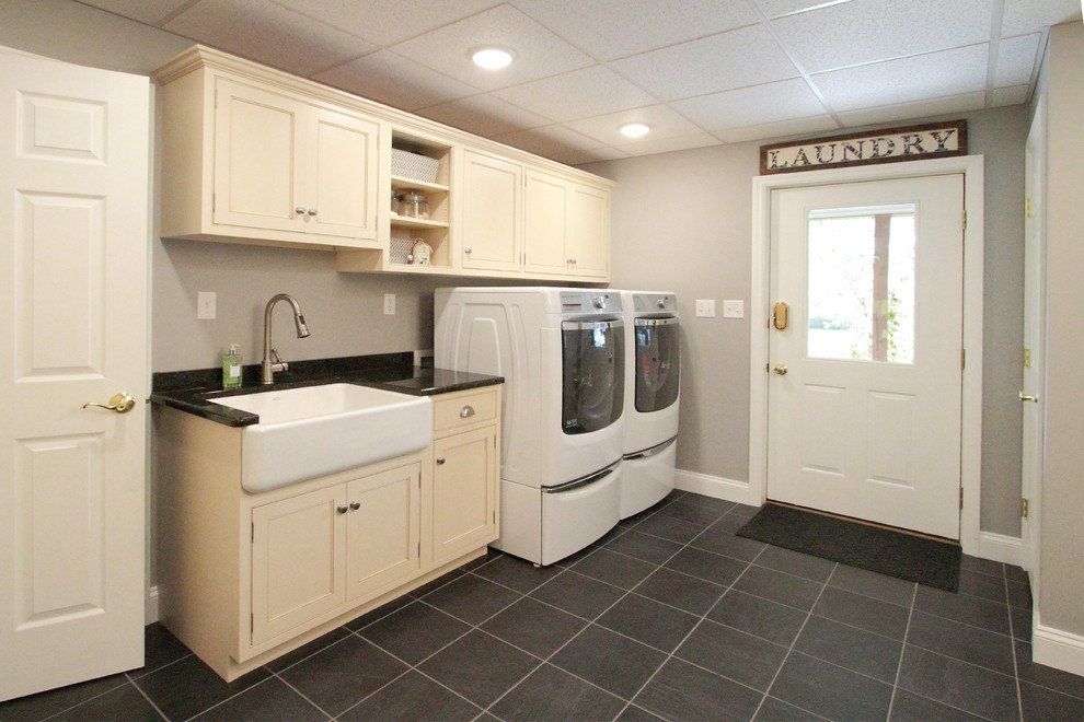 Photo of a mid-sized transitional single-wall utility room in Philadelphia with wood benchtops, porcelain floors, a farmhouse sink, shaker cabinets, grey walls, a side-by-side washer and dryer and beige cabinets.