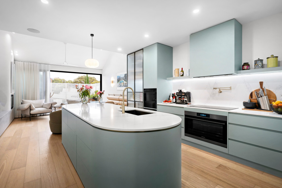 Photo of a contemporary galley kitchen in Melbourne with an undermount sink, flat-panel cabinets, turquoise cabinets, white splashback, stone slab splashback, black appliances, medium hardwood floors, with island, brown floor and white benchtop.