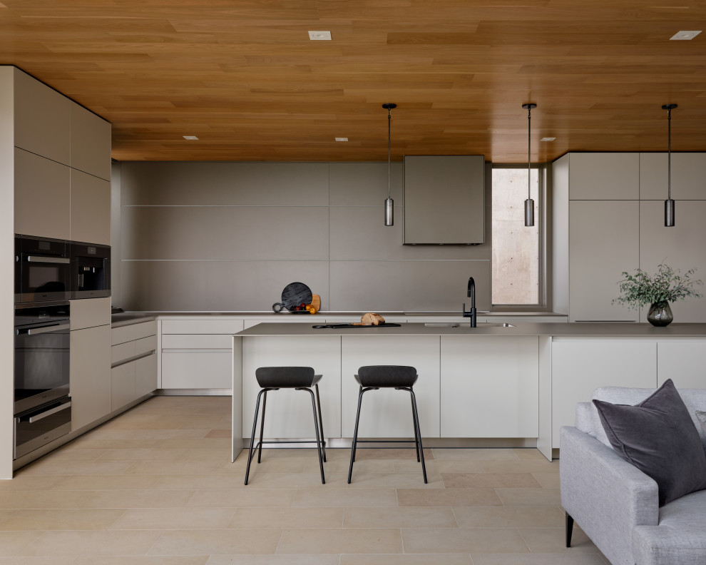 На фото: большая параллельная кухня-гостиная в стиле модернизм с врезной мойкой, плоскими фасадами, серыми фасадами, серым фартуком, полом из известняка, островом, бежевым полом и деревянным потолком с