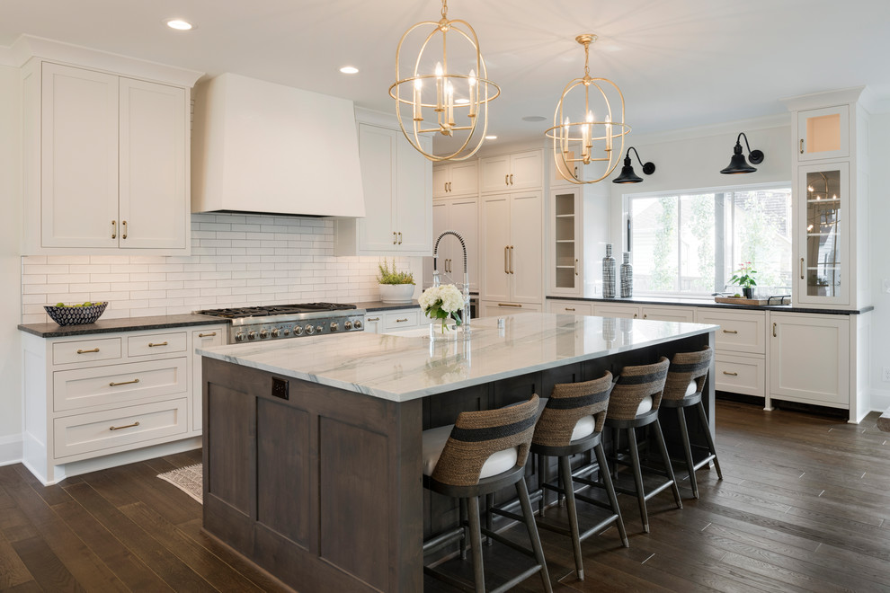 Photo of a large transitional l-shaped eat-in kitchen in Minneapolis with a farmhouse sink, shaker cabinets, white cabinets, marble benchtops, white splashback, ceramic splashback, stainless steel appliances, dark hardwood floors, with island, brown floor and grey benchtop.