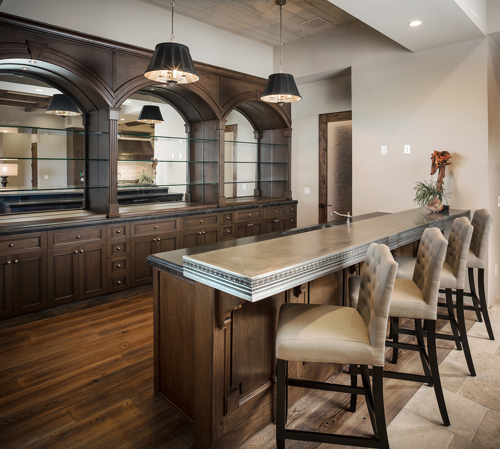 Inspiration for an expansive mediterranean galley wet bar in Phoenix with a drop-in sink, recessed-panel cabinets, dark wood cabinets, zinc benchtops, beige floor and grey benchtop.