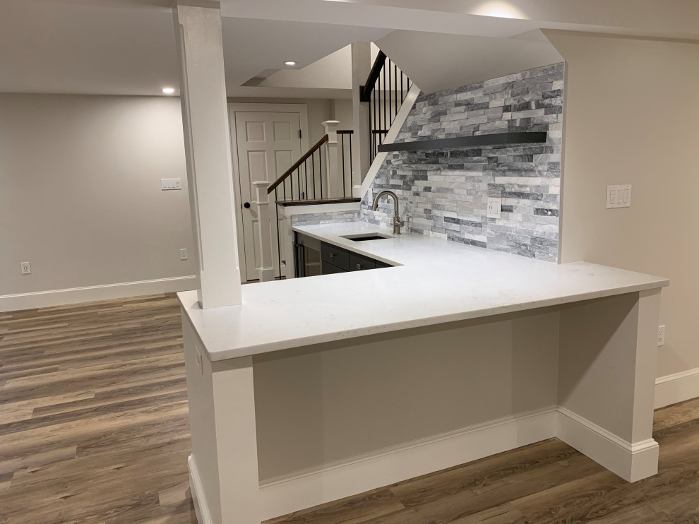 Mid-sized transitional l-shaped wet bar in Boston with an undermount sink, flat-panel cabinets, grey cabinets, solid surface benchtops, grey splashback, stone tile splashback, medium hardwood floors, brown floor and white benchtop.