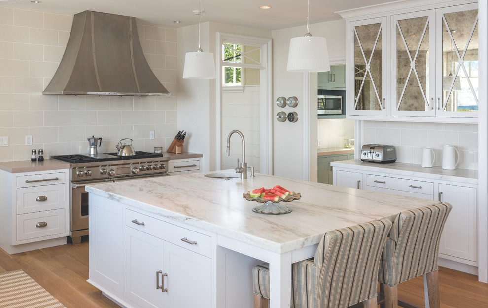 Large beach style u-shaped separate kitchen in Portland Maine with an undermount sink, white cabinets, beige splashback, stainless steel appliances, light hardwood floors, with island, shaker cabinets, marble benchtops, porcelain splashback and brown floor.