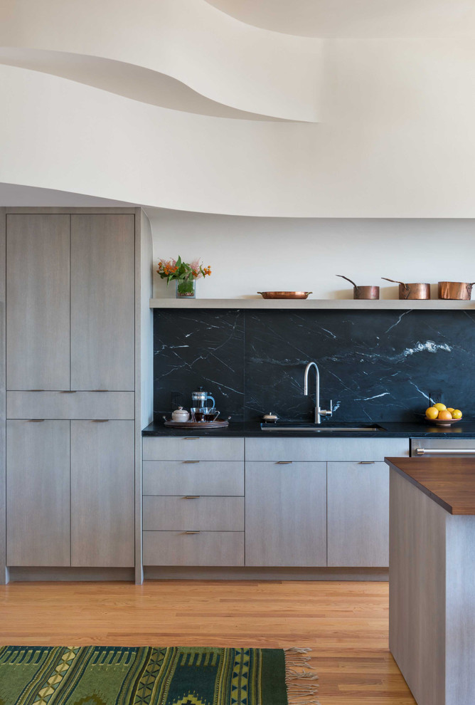 Mid-sized eclectic single-wall open plan kitchen in Boston with an undermount sink, flat-panel cabinets, light wood cabinets, soapstone benchtops, black splashback, stone slab splashback, panelled appliances, light hardwood floors and with island.