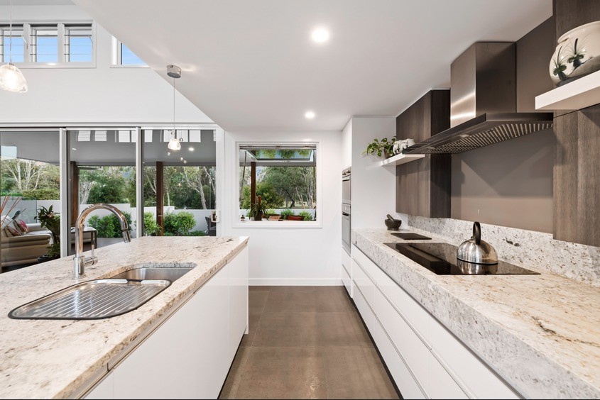 This is an example of a mid-sized beach style galley open plan kitchen in Sunshine Coast with a drop-in sink, recessed-panel cabinets, white cabinets, quartz benchtops, beige splashback, stone slab splashback, stainless steel appliances, cement tiles, with island, grey floor and beige benchtop.