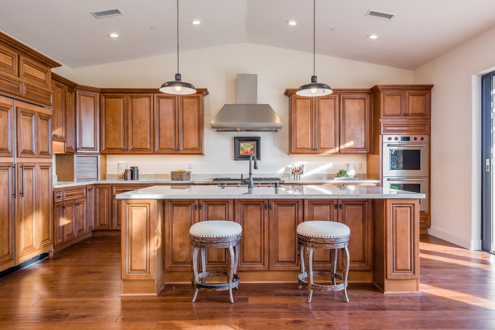 Design ideas for a traditional l-shaped kitchen in Santa Barbara with a farmhouse sink, raised-panel cabinets, medium wood cabinets, stainless steel appliances, medium hardwood floors, with island, brown floor and white benchtop.