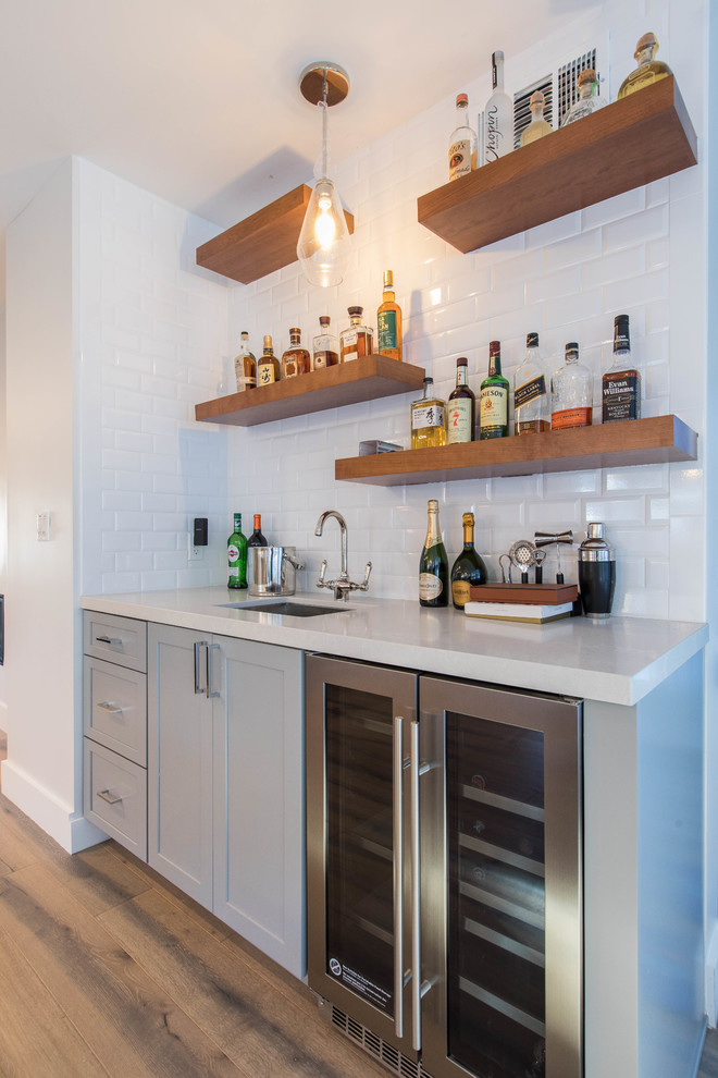 This is an example of a small transitional single-wall wet bar in Los Angeles with an undermount sink, shaker cabinets, grey cabinets, quartz benchtops, white splashback, subway tile splashback, light hardwood floors, beige floor and white benchtop.