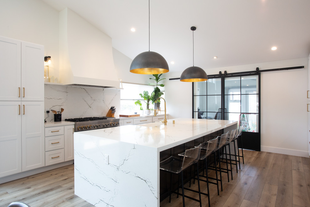 Kitchen - mid-sized modern single-wall brown floor kitchen idea in Orange County with shaker cabinets, white cabinets, white backsplash, stainless steel appliances, an island, white countertops and a farmhouse sink