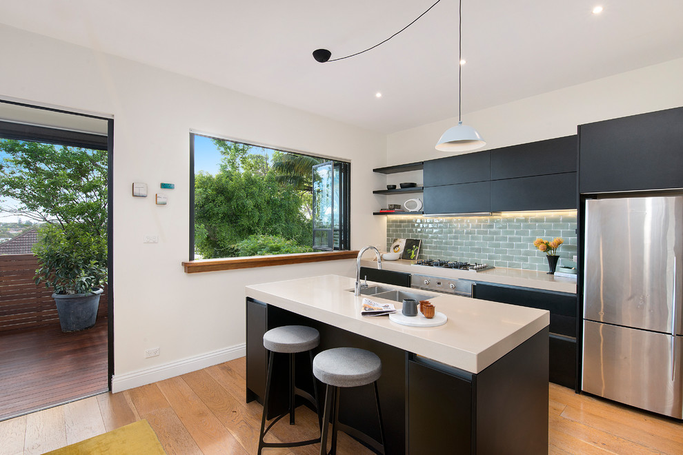 Photo of a contemporary single-wall kitchen in Sydney with a double-bowl sink, flat-panel cabinets, black cabinets, subway tile splashback, stainless steel appliances, medium hardwood floors, with island, brown floor, solid surface benchtops, grey splashback and white benchtop.
