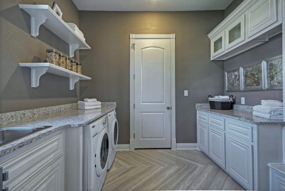 Photo of a large contemporary galley dedicated laundry room in Dallas with raised-panel cabinets, white cabinets, granite benchtops, grey walls, porcelain floors, a side-by-side washer and dryer and a drop-in sink.