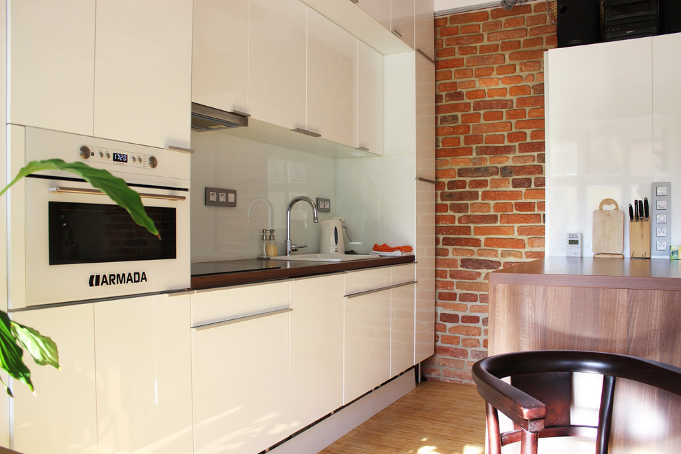 Photo of an industrial galley kitchen in Other with flat-panel cabinets, a drop-in sink, white cabinets, white splashback, white appliances and glass sheet splashback.