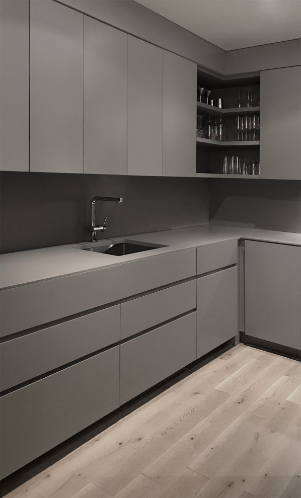 Mid-sized minimalist l-shaped medium tone wood floor and gray floor kitchen photo with an undermount sink, flat-panel cabinets, gray cabinets, solid surface countertops, gray backsplash, paneled appliances, no island and gray countertops