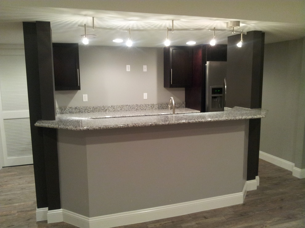 This is an example of an expansive contemporary single-wall wet bar in St Louis with shaker cabinets, dark wood cabinets, granite benchtops and light hardwood floors.