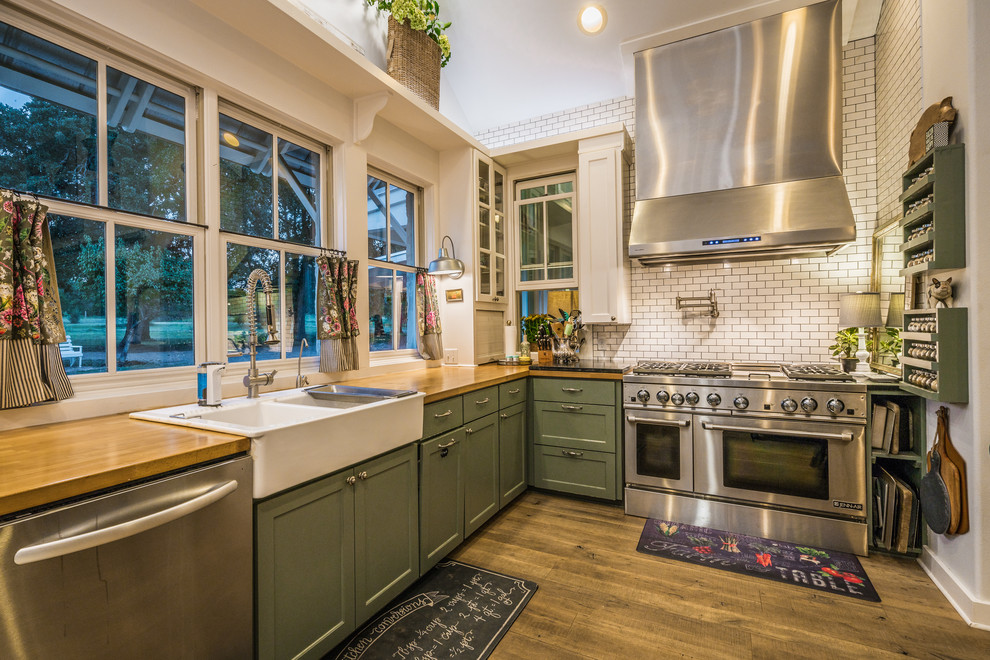 This is an example of a country l-shaped kitchen in Austin with a farmhouse sink, shaker cabinets, green cabinets, wood benchtops, white splashback, subway tile splashback, stainless steel appliances and medium hardwood floors.