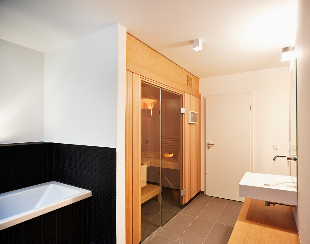 This is an example of a mid-sized contemporary bathroom in Frankfurt with a wall-mount sink, a drop-in tub, black tile, white walls, light wood cabinets, wood benchtops, mosaic tile and with a sauna.