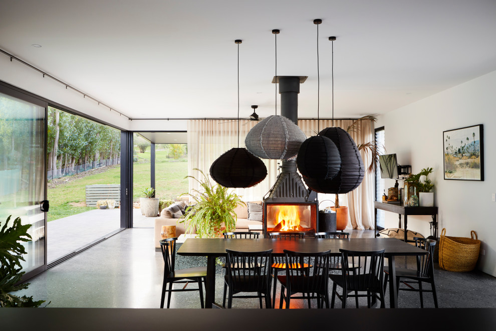Photo of a large contemporary dining room in Other with white walls, concrete floors, a two-sided fireplace, a concrete fireplace surround and black floor.