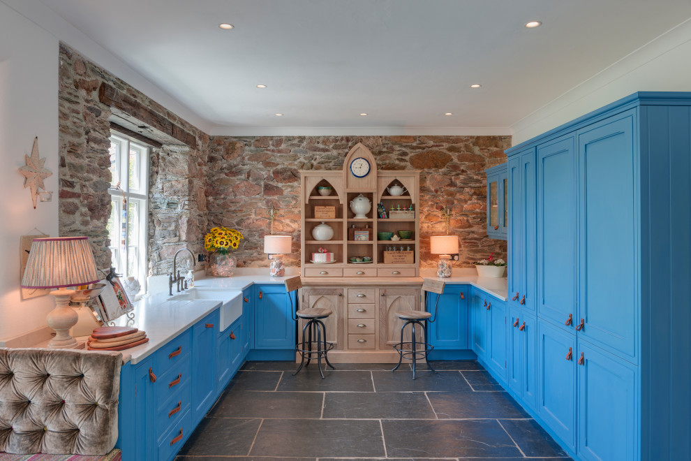 Photo of a large country u-shaped kitchen in Devon with a farmhouse sink, beaded inset cabinets, blue cabinets, solid surface benchtops, brown splashback, stone slab splashback, slate floors, no island, grey floor, white benchtop and panelled appliances.