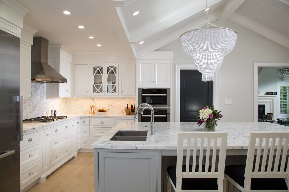 Mid-sized transitional l-shaped light wood floor kitchen photo with an undermount sink, recessed-panel cabinets, white cabinets, marble countertops, white backsplash, marble backsplash, stainless steel appliances and an island