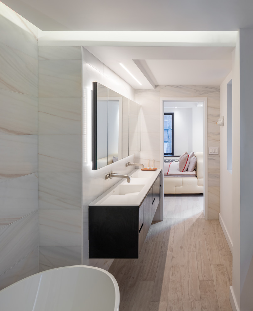 Photo of a large contemporary master bathroom in New York with flat-panel cabinets, black cabinets, a freestanding tub, an alcove shower, a wall-mount toilet, black tile, stone tile, black walls, light hardwood floors, an integrated sink, engineered quartz benchtops, beige floor and a hinged shower door.