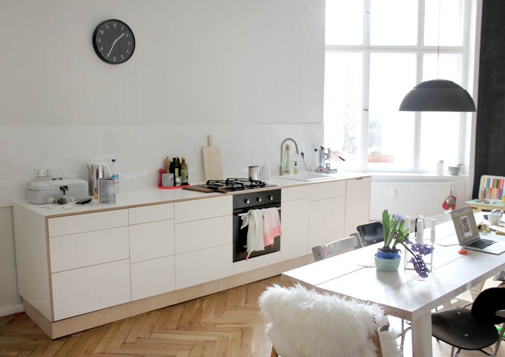 Inspiration for a mid-sized contemporary single-wall eat-in kitchen in Berlin with a drop-in sink, flat-panel cabinets, white cabinets, white splashback, glass tile splashback, black appliances, medium hardwood floors and no island.