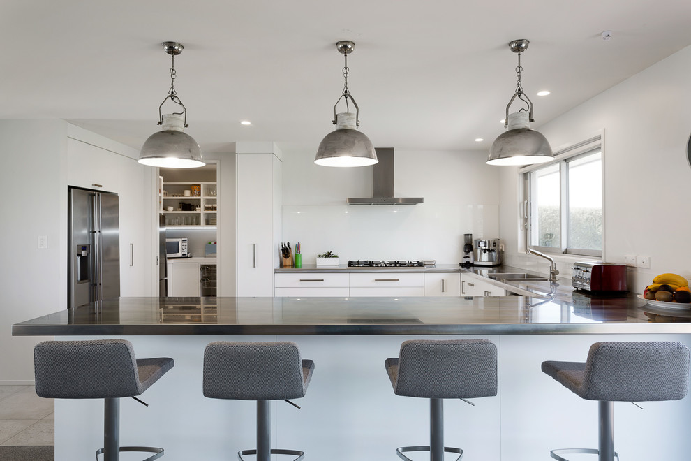 Design ideas for a large modern u-shaped open plan kitchen in Auckland with a double-bowl sink, flat-panel cabinets, white cabinets, stainless steel benchtops, white splashback, glass sheet splashback, stainless steel appliances, porcelain floors and a peninsula.