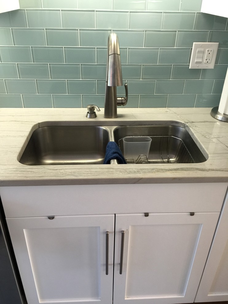 Medium tone wood floor kitchen photo with a double-bowl sink, flat-panel cabinets, white cabinets, quartzite countertops, blue backsplash, glass tile backsplash and stainless steel appliances