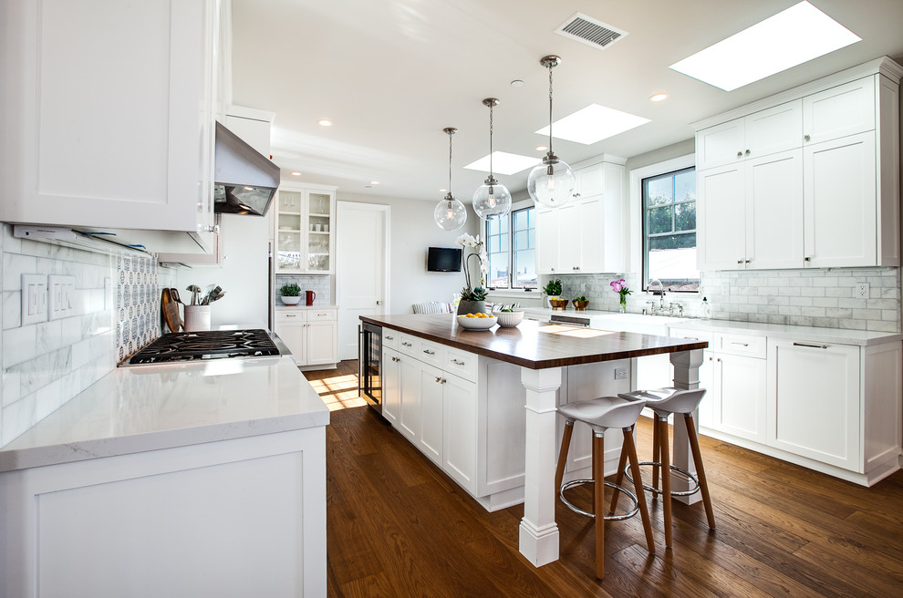This is an example of a large beach style u-shaped eat-in kitchen in Los Angeles with a farmhouse sink, shaker cabinets, white cabinets, marble benchtops, white splashback, marble splashback, stainless steel appliances, dark hardwood floors, with island, brown floor and white benchtop.