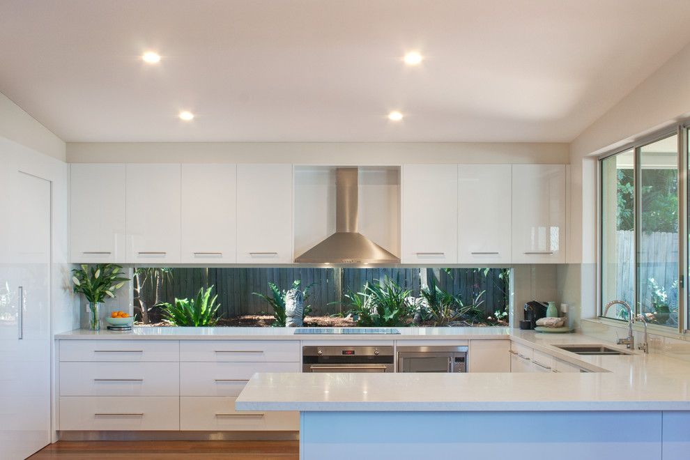 Design ideas for a mid-sized contemporary u-shaped open plan kitchen in Sydney with an undermount sink, flat-panel cabinets, white cabinets, quartz benchtops, window splashback, stainless steel appliances, medium hardwood floors and white benchtop.
