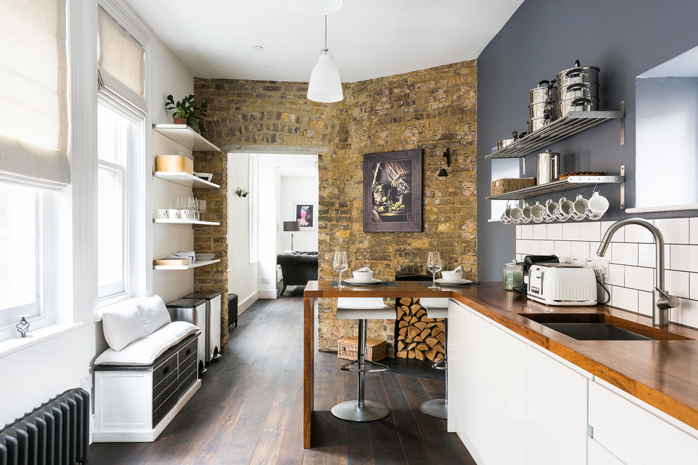 This is an example of a country l-shaped kitchen in London with an undermount sink, flat-panel cabinets, white cabinets, wood benchtops, white splashback, subway tile splashback, dark hardwood floors, a peninsula and brown floor.