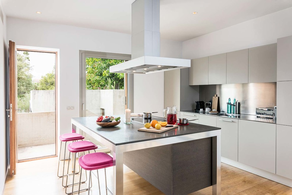 Contemporary kitchen in Palma de Mallorca with a single-bowl sink, flat-panel cabinets, grey cabinets, stainless steel benchtops, metallic splashback, light hardwood floors and with island.