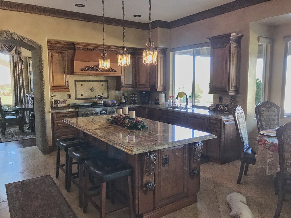 This is an example of a large traditional l-shaped open plan kitchen in Phoenix with a farmhouse sink, raised-panel cabinets, medium wood cabinets, granite benchtops, beige splashback, travertine splashback, stainless steel appliances, travertine floors, with island, beige floor and beige benchtop.