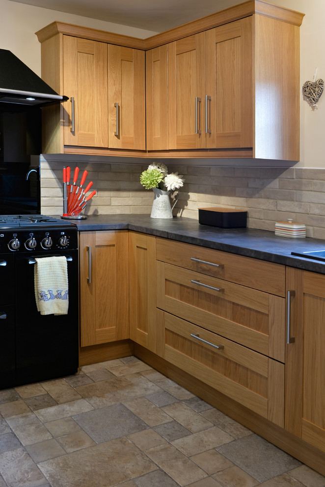 Photo of a mid-sized modern l-shaped separate kitchen in Cornwall with a drop-in sink, shaker cabinets, brown cabinets, laminate benchtops, black splashback, glass sheet splashback, black appliances, laminate floors, no island, beige floor and black benchtop.