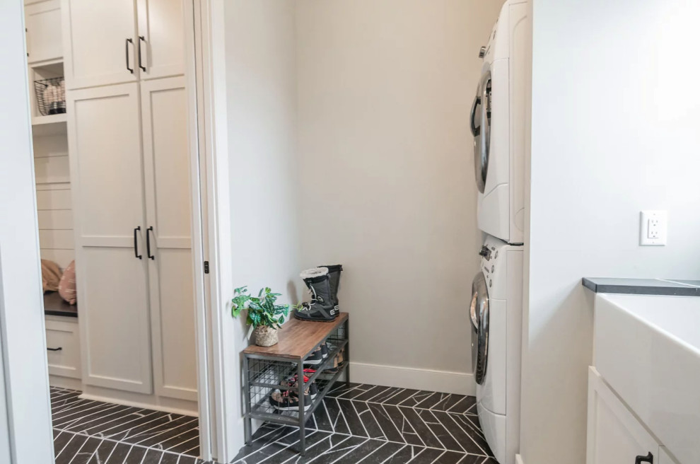Medium sized classic galley utility room in Minneapolis with a belfast sink, shaker cabinets, white cabinets, engineered stone countertops, white splashback, ceramic splashback, grey walls, ceramic flooring, a stacked washer and dryer, black floors and black worktops.