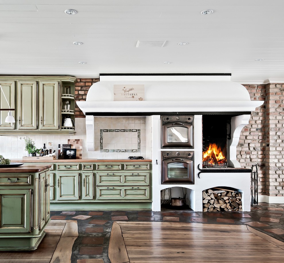 This is an example of a large country eat-in kitchen in Gothenburg with green cabinets, wood benchtops, multi-coloured splashback, with island, multi-coloured floor and raised-panel cabinets.