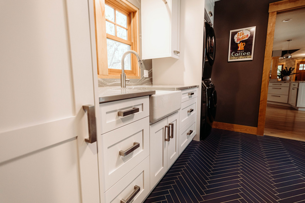 Photo of a large arts and crafts galley utility room in Other with a farmhouse sink, shaker cabinets, white cabinets, quartz benchtops, grey splashback, engineered quartz splashback, grey walls, porcelain floors, a stacked washer and dryer, blue floor and grey benchtop.