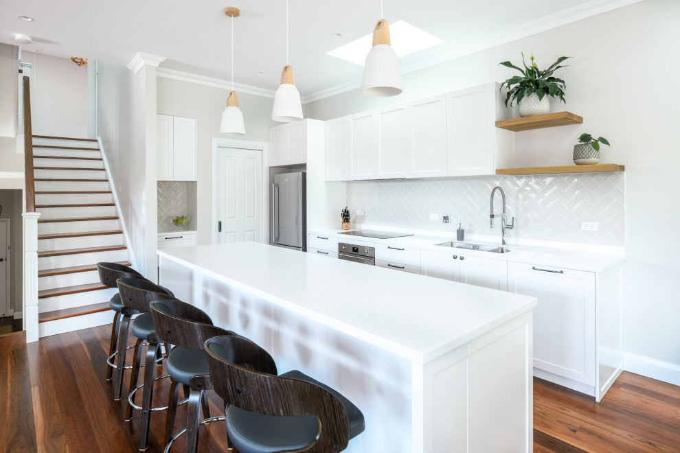 Large contemporary galley open plan kitchen in Sydney with an undermount sink, shaker cabinets, white cabinets, solid surface benchtops, beige splashback, porcelain splashback, stainless steel appliances, dark hardwood floors, with island, brown floor and white benchtop.
