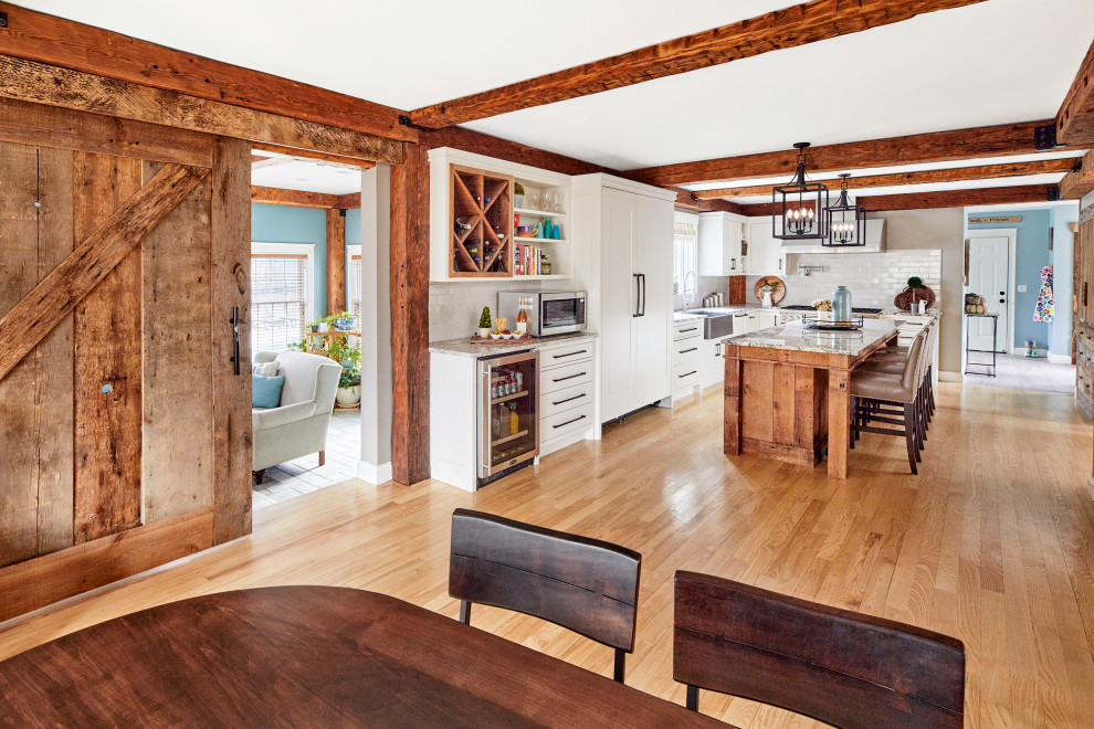 This is an example of a large rural l-shaped kitchen/diner in Portland Maine with a belfast sink, shaker cabinets, white cabinets, white splashback, metro tiled splashback, integrated appliances, light hardwood flooring, an island, multicoloured worktops, exposed beams and granite worktops.