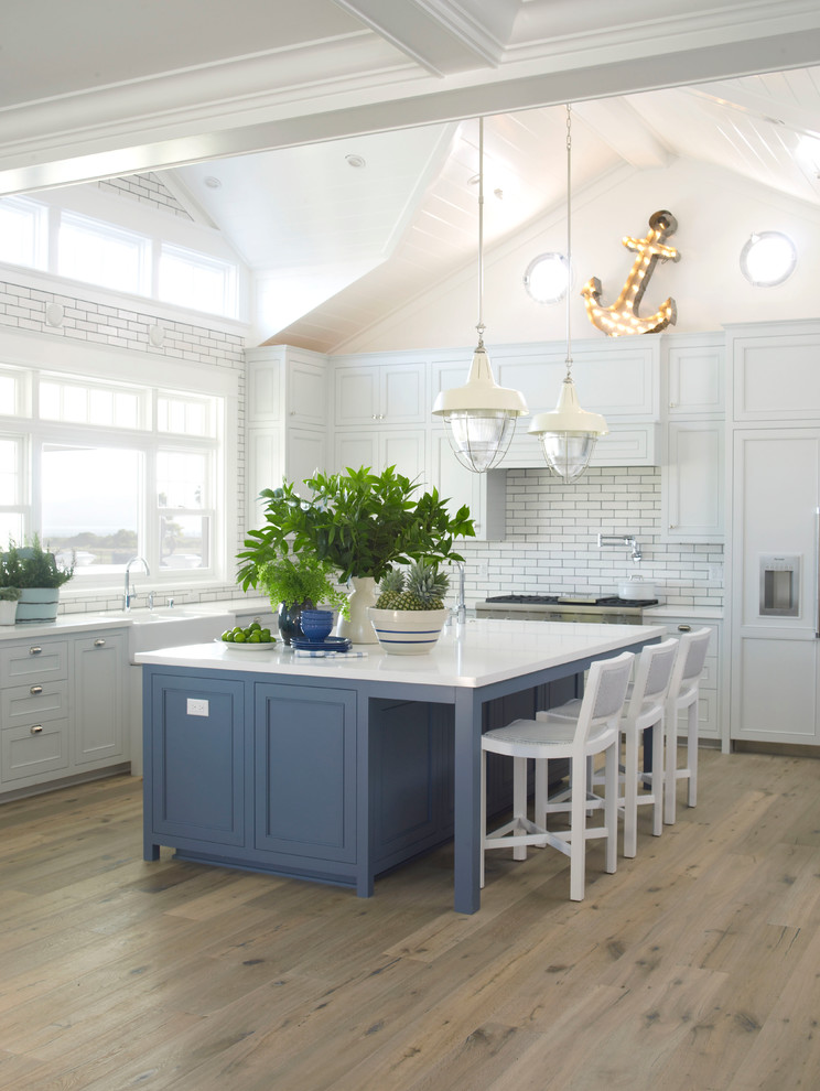 Design ideas for a large beach style l-shaped kitchen in San Diego with a farmhouse sink, shaker cabinets, blue cabinets, quartz benchtops, white splashback, panelled appliances, light hardwood floors, with island and subway tile splashback.