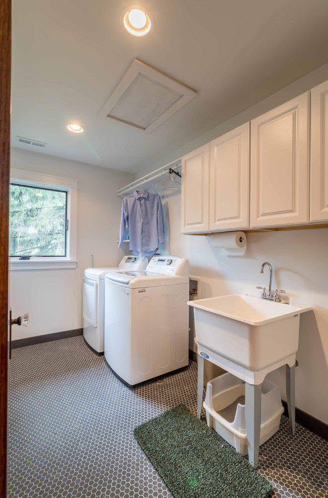 Photo of a mid-sized arts and crafts galley dedicated laundry room in Chicago with an utility sink, raised-panel cabinets, white cabinets, white walls, ceramic floors, a side-by-side washer and dryer, grey floor, white splashback, cement tile splashback, wallpaper, wallpaper and white benchtop.