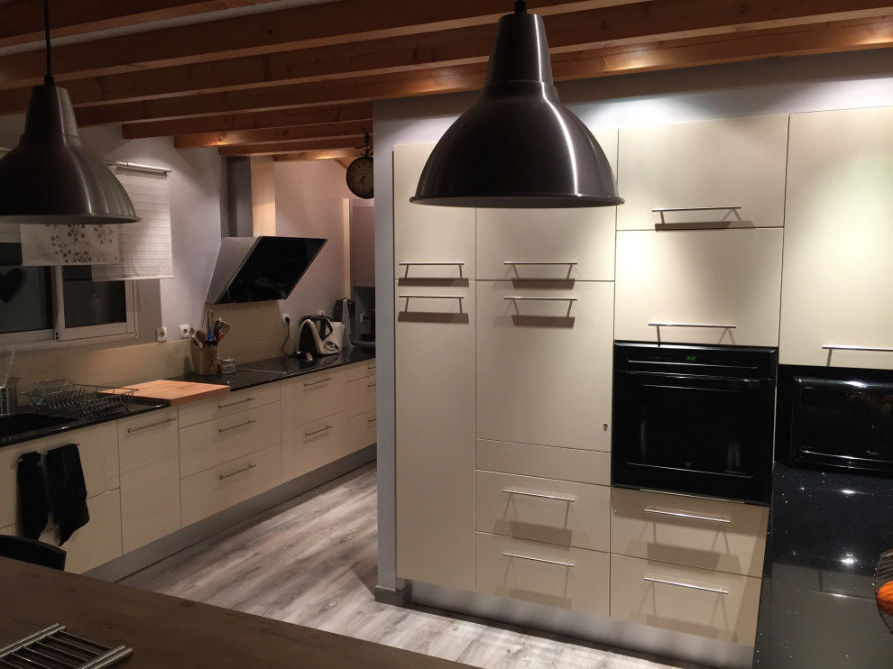 This is an example of a large industrial l-shaped open plan kitchen in Toulouse with an integrated sink, beaded inset cabinets, beige cabinets, quartzite benchtops, beige splashback, glass tile splashback, panelled appliances, light hardwood floors, a peninsula, grey floor, black benchtop and exposed beam.