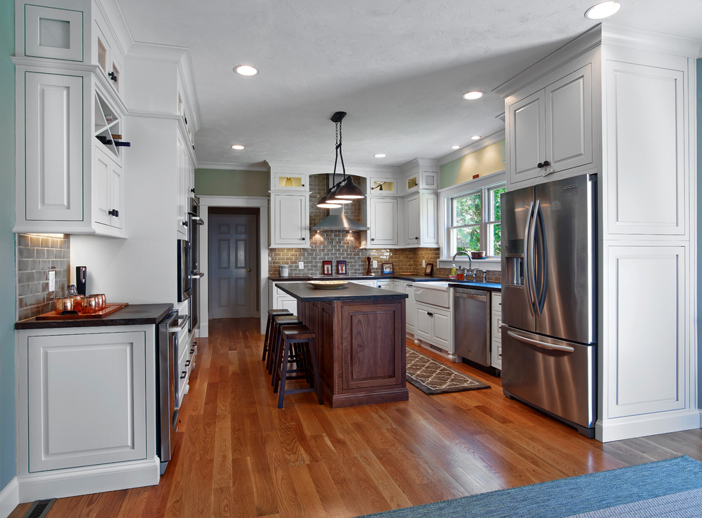Example of a mid-sized transitional u-shaped medium tone wood floor and brown floor enclosed kitchen design in Providence with a farmhouse sink, raised-panel cabinets, white cabinets, gray backsplash, subway tile backsplash, stainless steel appliances and an island
