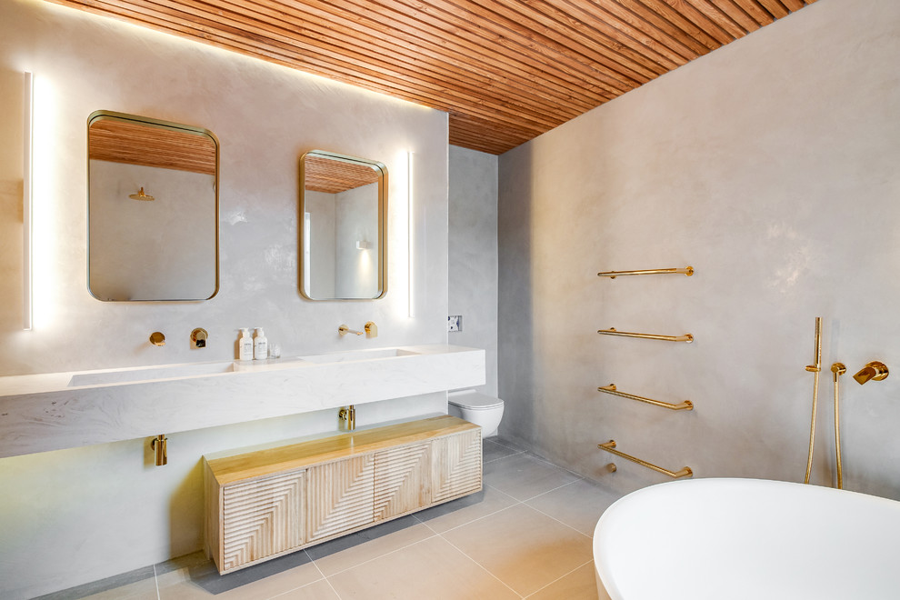 Photo of a contemporary bathroom in Other with light wood cabinets, a freestanding tub, a wall-mount toilet, grey walls, an integrated sink, beige floor and white benchtops.