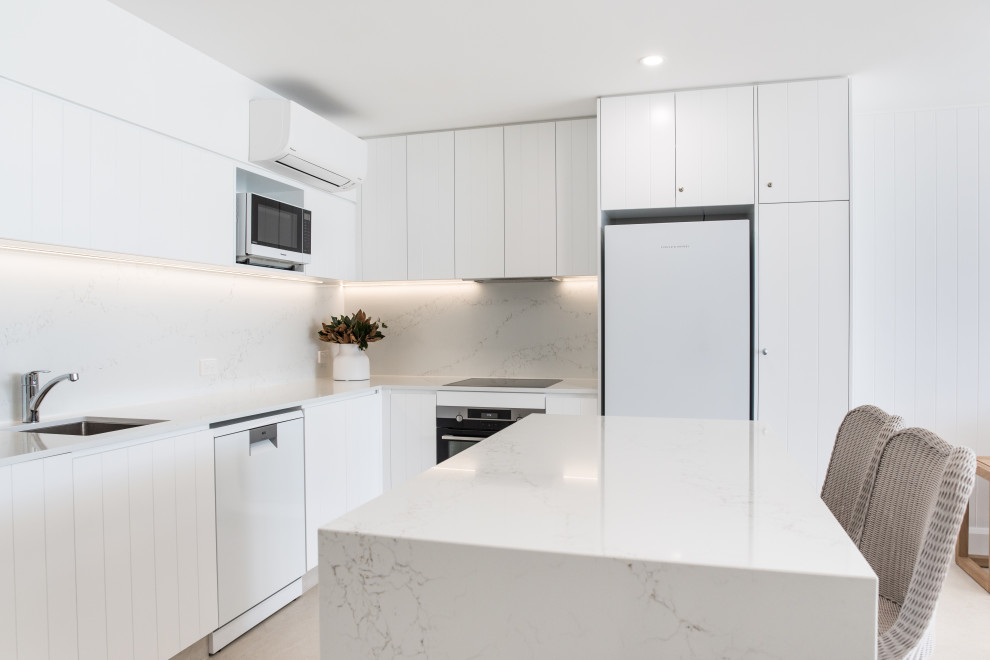 This is an example of a small beach style l-shaped open plan kitchen in Sunshine Coast with quartz benchtops, white splashback, engineered quartz splashback, stainless steel appliances, porcelain floors, with island, beige floor and white benchtop.