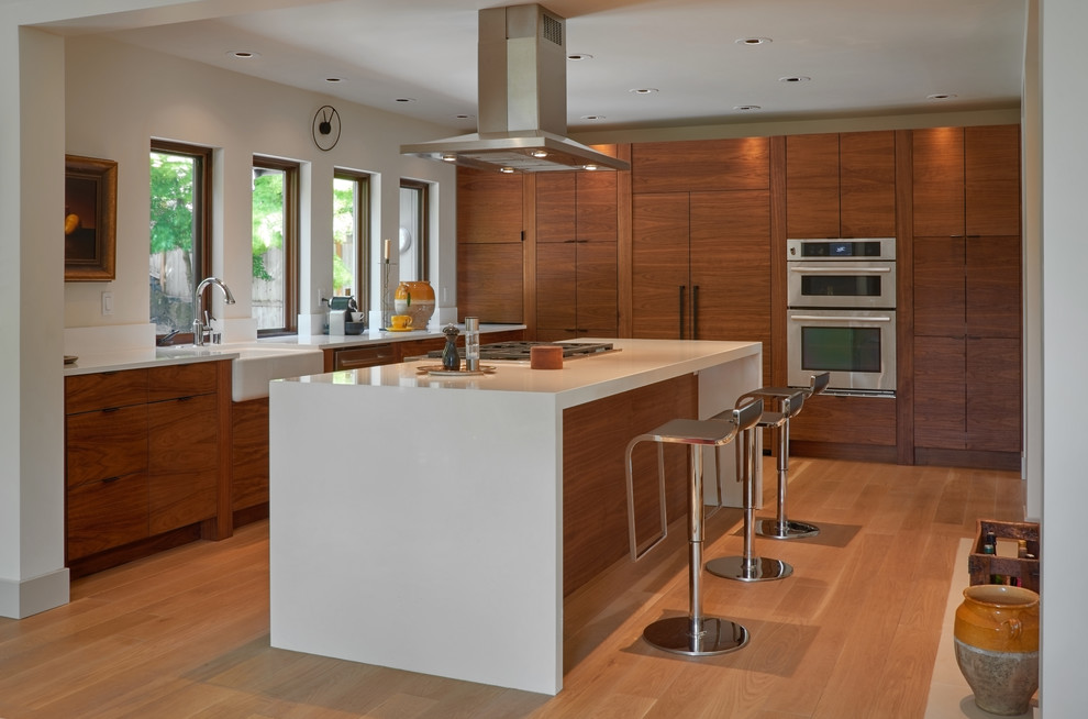 This is an example of a mid-sized midcentury l-shaped eat-in kitchen in Seattle with a farmhouse sink, flat-panel cabinets, medium wood cabinets, quartz benchtops, white splashback, stainless steel appliances, light hardwood floors and with island.