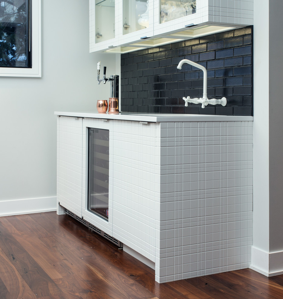 Photo of a mid-sized contemporary single-wall wet bar in Other with an undermount sink, flat-panel cabinets, white cabinets, solid surface benchtops, black splashback, subway tile splashback, dark hardwood floors and brown floor.